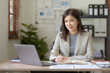 Beautiful Asian businesswoman using her laptop enjoying work, taking notes, reviewing work done.