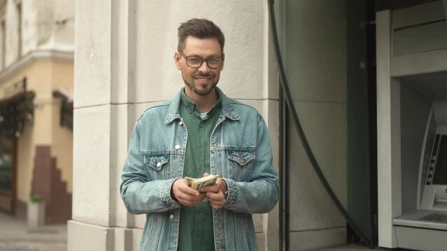 Portrait of cheerful caucasian rich man counting money cashed out from bank credit card using ATM terminal on street. Beautiful happy man looking at camera smiling after financial operations.