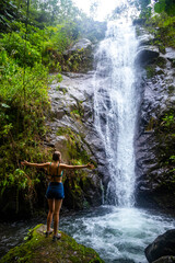 A beautiful girl spreads her arms while standing under a tropical waterfall in Costa Rica; swimming in a hidden waterfall in the rainforest