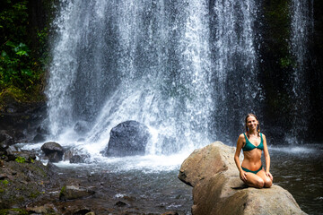 Naklejka na ściany i meble beautiful girl in a bikini sitting under a tropical waterfall in Costa Rica; swimming in a hidden waterfall in the rainforest