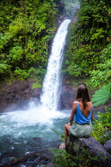 A beautiful girl sits on rocks under a powerful tropical waterfall in Costa Rica; la paz waterfall in the jungle