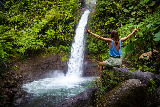 A beautiful girl sits on rocks under a powerful tropical waterfall in Costa Rica; la paz waterfall in the jungle