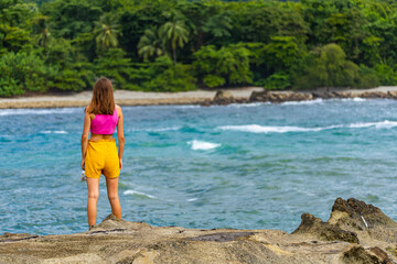 Fototapeta premium A beautiful girl in colorful clothing stands on the rocks admiring a tropical beach with palm trees; paradise beach in Costa Rica