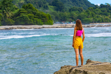 A beautiful girl in colorful clothing stands on the rocks admiring a tropical beach with palm trees; paradise beach in Costa Rica