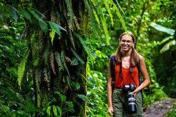 Naklejka na ściany i meble backpacker girl with camera walks through dense jungle in monteverde cloud forest, Costa Rica; walk through fairy tale, magical tropical rainforest; wild nature of Costa Rica