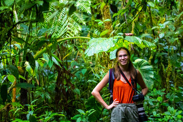 backpacker girl with camera stands under large leaves in monteverde cloud forest, Costa Rica; walk through fairy tale, magical tropical rainforest; wild nature of Costa Rica