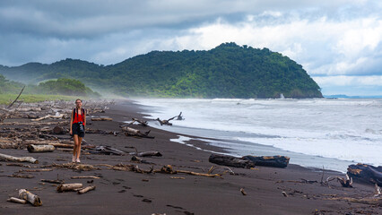girl walking along Costa Rica's black sand beach during a storm; large tree trunks tossed onto the beach by a hurricane