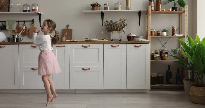 Funny little girl wear festive fluffy pink skirt jumping, dancing, moving barefoot in cozy warm fashionable kitchen, looking carefree, feel happy, kid fooling around alone at home. Active child, fun