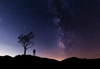 Hiker silhouette stands near the tree on the hill, behind them fantastic beautiful night sky with milky way galaxy. © Inga Av