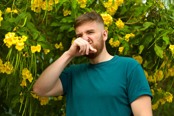 Portrait of handsome young allergic man is suffering from pollen allergy or cold on natural flowers, flowering tree background at spring or sunny summer day, sneezes, blowing his runny nose rubs eyes