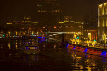 Fototapeta na wymiar City embankment decorated with gold colored illuminated tree shaped artificial Christmas decorations at night. Tourboat on water. Selective focus. Winter holidays theme.