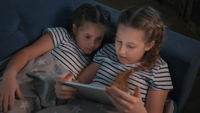 Two Cute Girls Lying On Couch Under Warm Blanket In Room Are Looking At Screen Of Digital Tablet.Modern Digital Entertainment Technologies For Children. Children Learn Through Digital Tablet, Have Fun