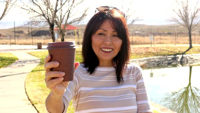 Smiling woman offers coffee to camera in park, sunglasses on head