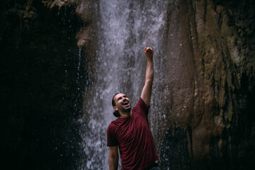 A young, joyful male tourist stands with his hand raised in a victorious gesture near a tropical waterfall among the rocks.