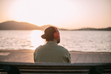 Fototapeta premium A young man is sitting on a bench with headphones on at sunset by the sea.