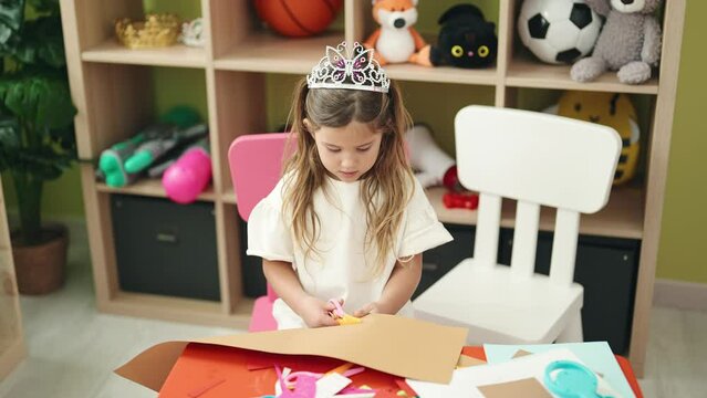 Adorable blonde girl student wearing princess crown cutting paper at kindergarten