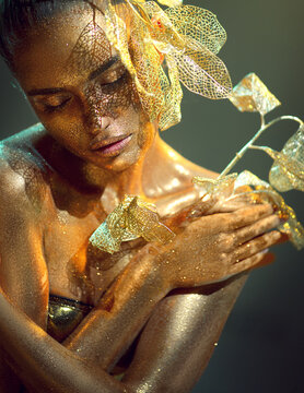High Fashion gold model woman with bright golden sparkles on skin, fantasy flower, portrait of beautiful girl glowing make-up. Art design gold dust sequins make up. Glitter glowing skin, jewellery.
