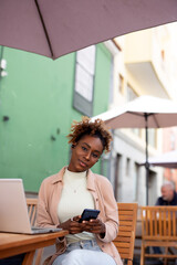 Happy Afro woman student on street with her laptop and using her mobile