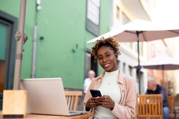 Happy Afro woman student on street with her laptop and using her mobile