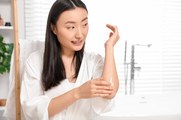 Beautiful young Asian woman applying body cream on elbow in bathroom, space for text