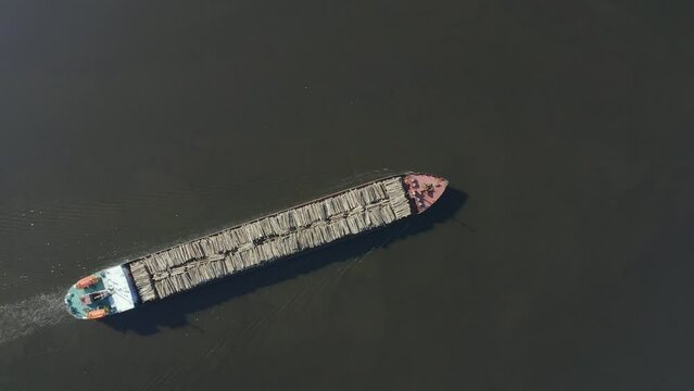 Long barge ship loaded with timber trunks of felled trees floats in the bay aerial top view