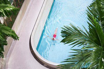 Vacation concept. Top view of slim young woman in red swimsuit swimming in the tropical pool.