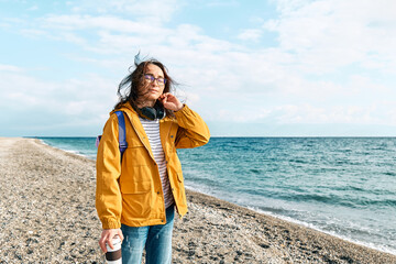 Smiling woman in yellow jacket with backpack and coffee mug portable walking along sea coast and enjoying the sunny day. Concept of hiking and activity in the low season.