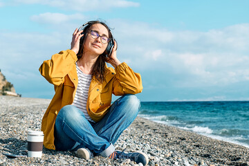 Woman with closed eyes listening music or podcast from smartphone application in headphones while sits on winter beach. Smiling middle-aged female in headphones listening music near the sea.
