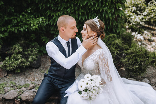 A stylish, young groom and a beautiful bride in a white dress with a bouquet in their hands, a diadem on their heads, sit in a park in nature. Wedding photo of smiling newlyweds.