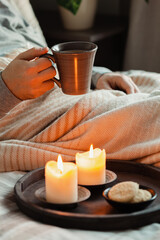 Fototapeta na wymiar A caucasian man relaxing at home, lighting candle, drinking coffee in bed under blanket