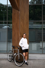 Fototapeta na wymiar Stylish african american woman in hoodie and shorts standing near bike on street 