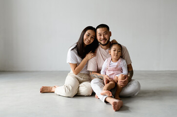 Joyful young asian family with baby daughter hugging and looking at camera while sitting on grey background