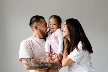 Happy young asian parents looking at each other while holding toddler daughter isolated on grey