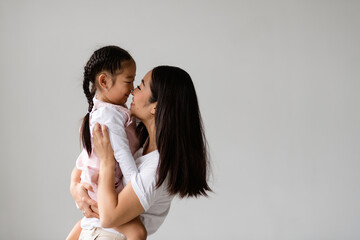 Happy young asian mother touching nose of toddler daughter while playing isolated on grey