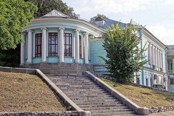 facade of a blue old building with white stone columns and red windows on a city street among green vegetation near a gray staircase