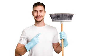 Young man holding a broom to clean his house cut out isolated smiling and pointing aside, showing...