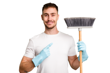Young man holding a broom to clean his house cut out isolated smiling and pointing aside, showing something at blank space.