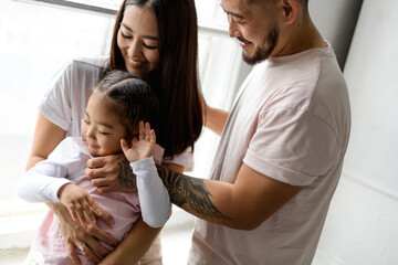 Cheerful young asian parents hugging toddler daughter near window indoors