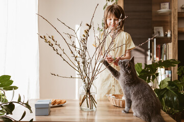 A happy girl hangs Easter eggs on a willow bouquet at home with a little kitten. Funny pets. Easter...