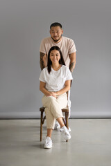 Smiling asian man looking at camera near brunete wife sitting on chair on grey background 