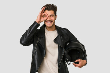 Young caucasian man holding a motrbike black helmet isolated excited keeping ok gesture on eye.