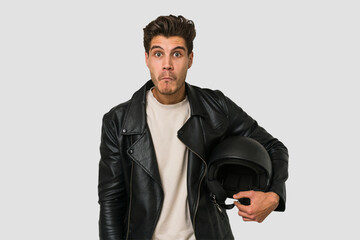 Young caucasian man holding a motrbike black helmet isolated shrugs shoulders and open eyes confused.
