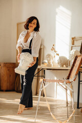 a brunette in a white shirt and trousers with suspenders in the artist's studio.