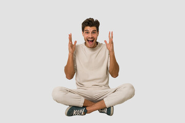 Young caucasian man sitting on the floor isolated on white background celebrating a victory or success, he is surprised and shocked.