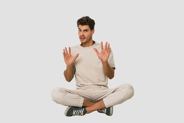 Young caucasian man sitting on the floor isolated on white background rejecting someone showing a...