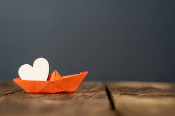 close-up of an orange miniature origami ship with a wooden heart on wooden table. dark background....