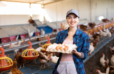 Happy successful female owner of poultry farm engaged in breeding of laying hens standing in chicken coop, holding carton tray of fresh organic eggs © JackF