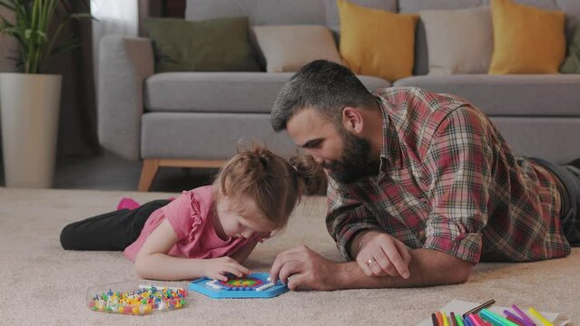 Young handsome father and cute daughter playing together. Dad and child making a puzzle with mosaic pegboard lying on the carpet at home. Educational toys for children. Home leisure. Father's day.