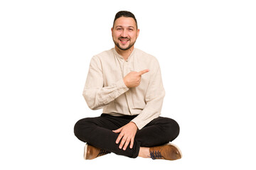 Adult latin man sitting on the floor cut out isolated smiling and pointing aside, showing something...