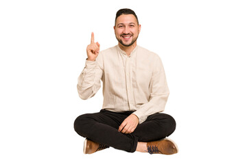 Adult latin man sitting on the floor cut out isolated showing number one with finger.